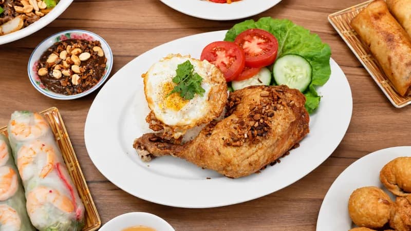 A Cadao Express spread on a wood table: grilled chicken rice platter with a fried egg, fresh shrimp spring rolls, peanut sauce, egg rolls and fried wontons